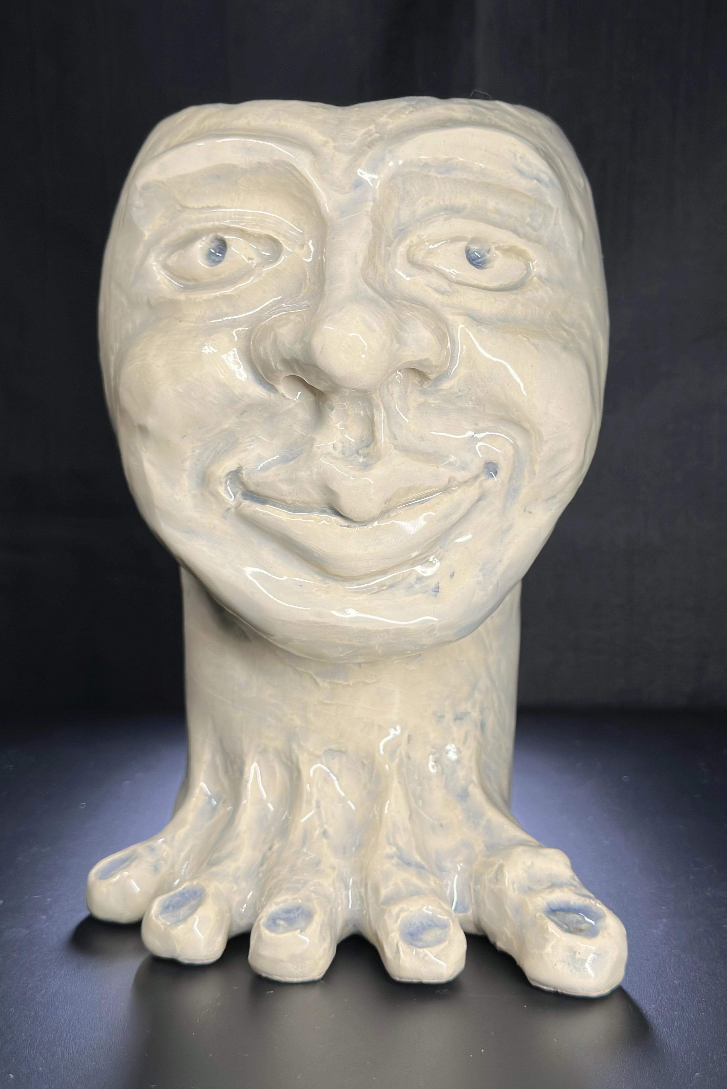 John: a glossy cream-colored ceramic bust with a broad smiling face, pale blue eyes, a long neck, and multiple rounded toe-like forms spreading from its base.