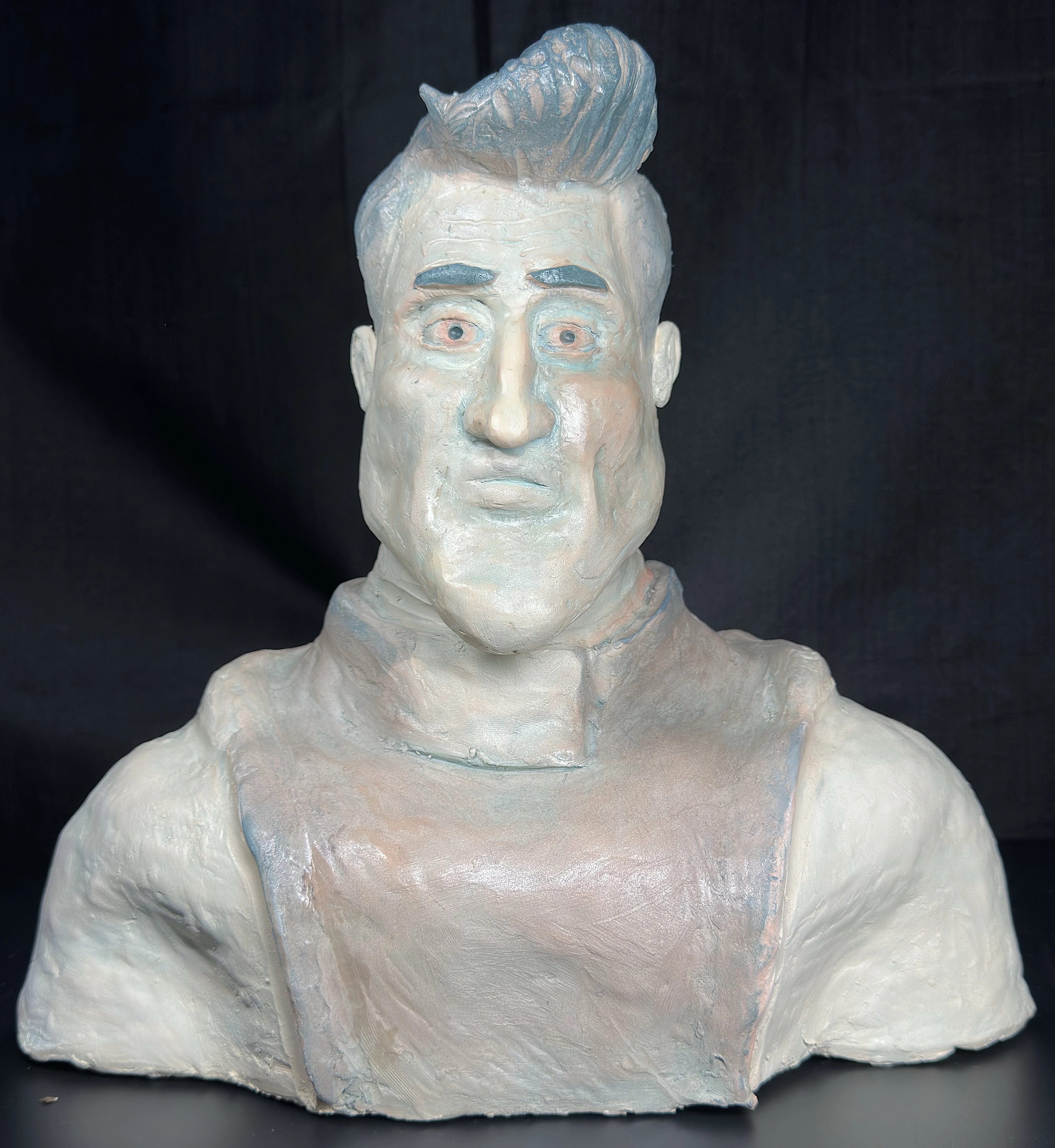 Metroman Bust: a pale blue-and-cream glazed ceramic bust with swept-up hair, blue eyebrows and eyes, broad shoulders, and a muscular upper body against a black backdrop.