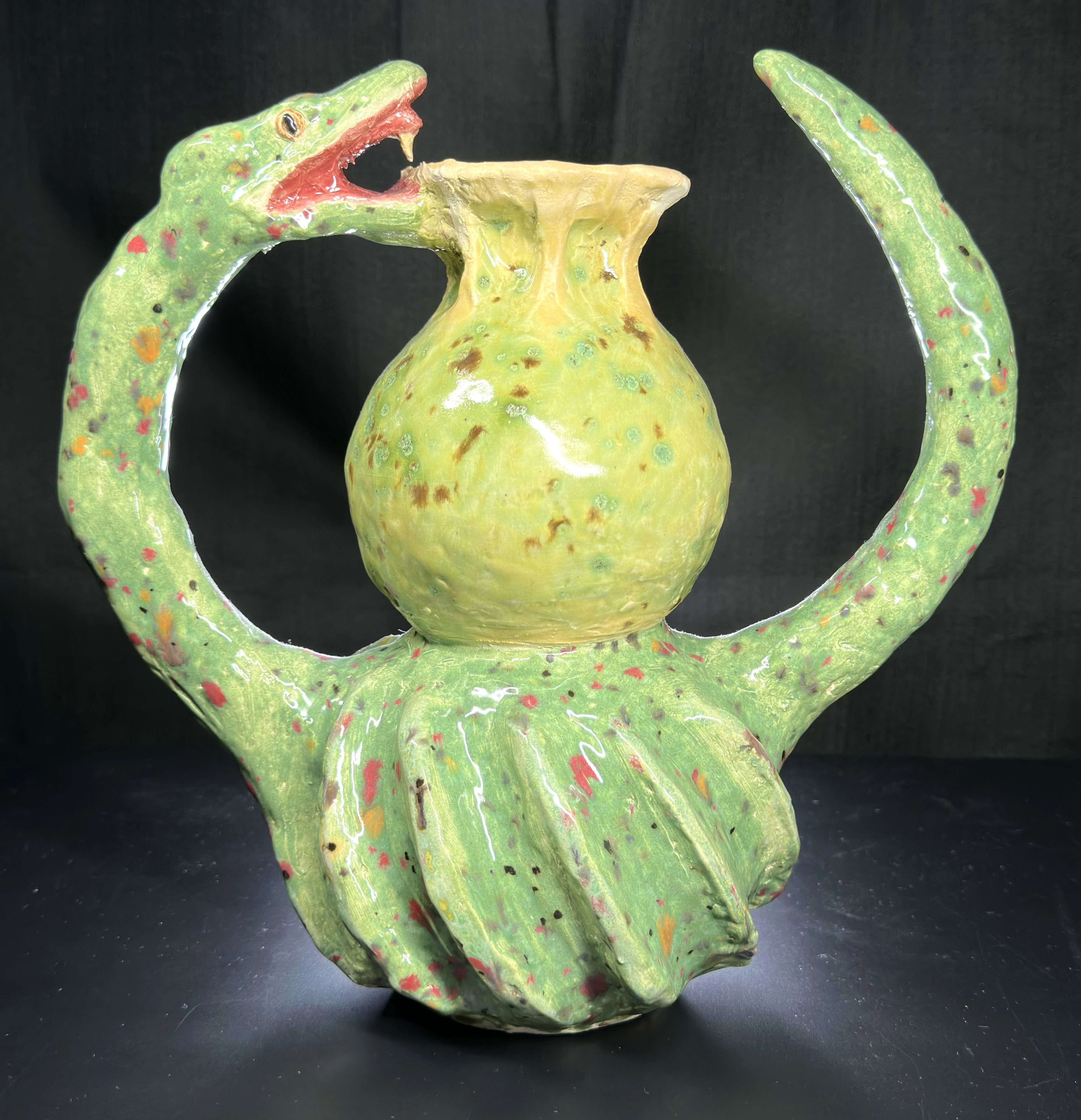 Snake Pot: a green and yellow glazed ceramic vessel encircled by a snake-like form, with an open red mouth on the left, a raised tail on the right, and red and brown speckles across the surface.