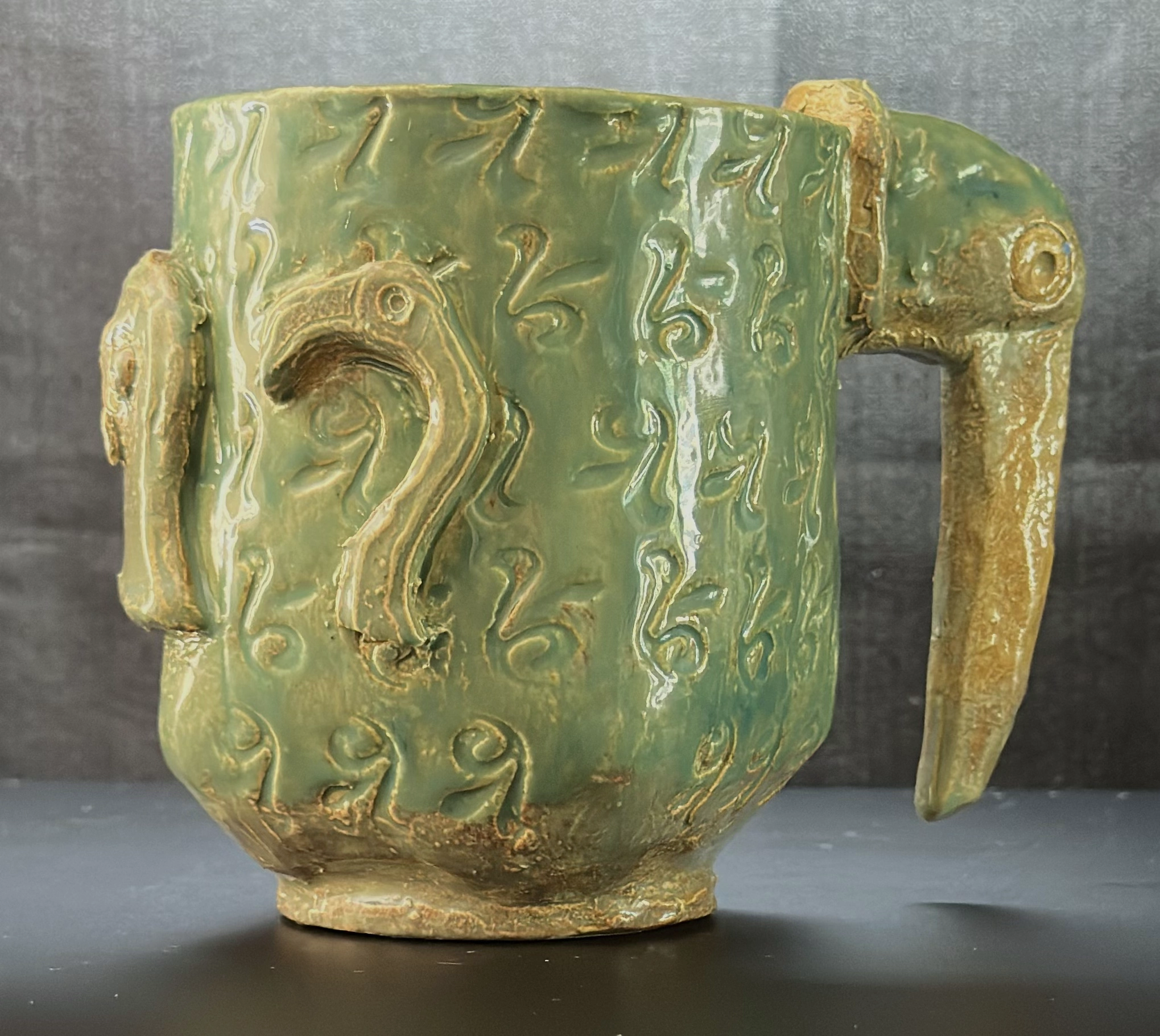 Tropical Bird Mug: a green glazed ceramic mug decorated with raised bird forms, including a parrot-like bird on the side and a bird head forming the handle on the right.